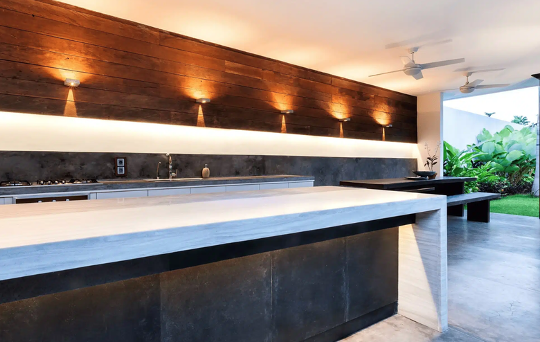 A modern outdoor kitchen with sleek stainless steel appliances, complemented by a stone seating area, surrounded by lush greenery and trees for a natural, inviting ambiance.