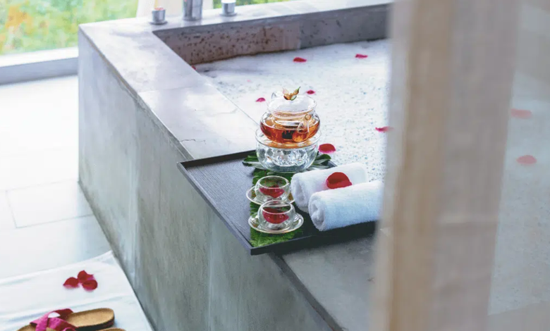 A serene spa setup featuring a stone bathtub adorned with rose petals, accompanied by a tray holding herbal tea, towels, and glass cups for a relaxing experience.