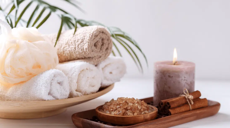This image depicts a serene spa setup with rolled towels, a lit candle, bath salts, cinnamon sticks, and a natural sponge, evoking a calming and relaxing ambiance.