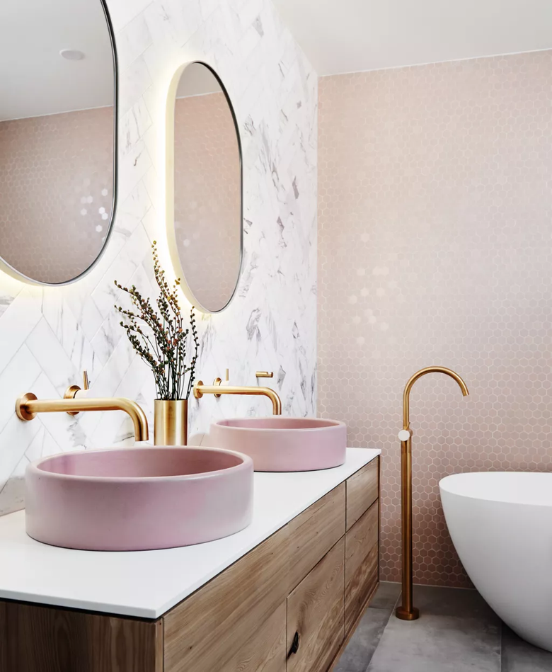 This stylish bathroom features soft pink vessel sinks with gold faucets, a freestanding bathtub, warm wood cabinetry, and a mix of marble and hexagonal tiled walls, creating a modern yet inviting ambiance.