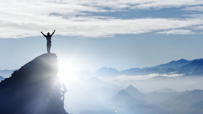 Eine Person, die auf einem Berggipfel steht und die Arme triumphierend in die Luft streckt, mit einer majestätischen Berglandschaft um sich.