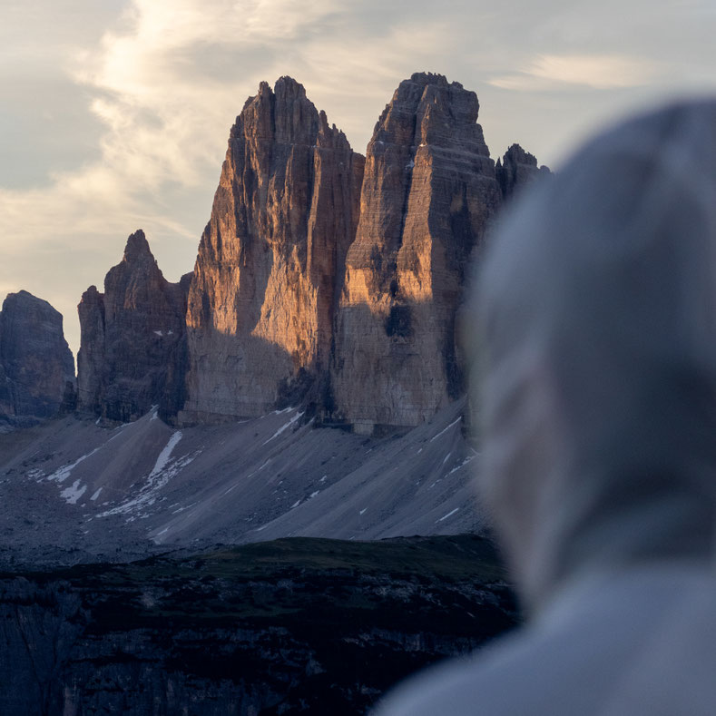 Die Drei Zinnen in den Dolomiten leuchten im warmen Licht der aufgehenden Sonne. Im unscharfen Vordergrund blickt eine Person in heller Outdoor-Kleidung auf die markanten Felsgipfel und genießt die ruhige Morgenstimmung.