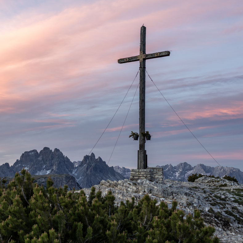 Ein schlichtes Gipfelkreuz aus Holz steht auf einem felsigen Bergplateau der Dolomiten. Im Hintergrund zeichnen sich markante Berggipfel unter einem Himmel in zarten Rosa-, Blau- und Violetttönen ab und verleihen der Szene eine ruhige Stimmung in der Morg