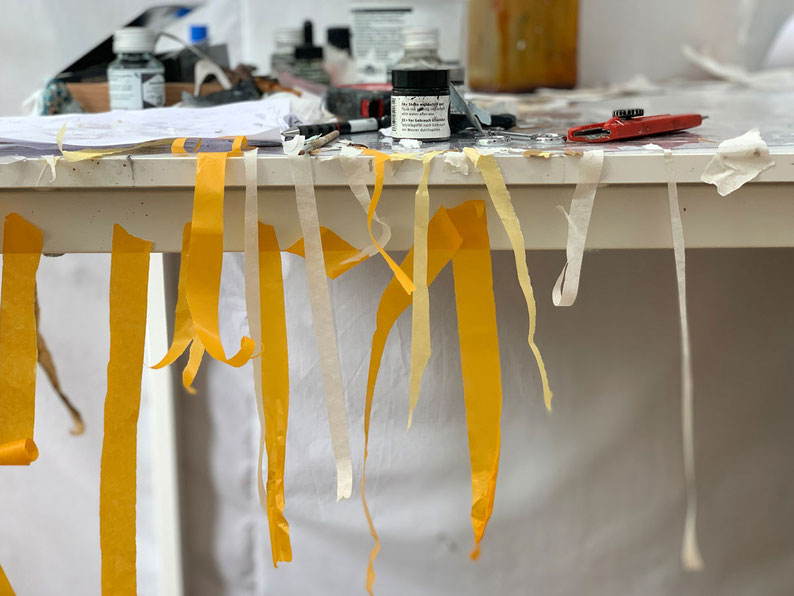 Arbeitstisch im Kunstatelier mit Tintenfass, Cutter, Stiften und herabhängenden gelben Klebbändern.