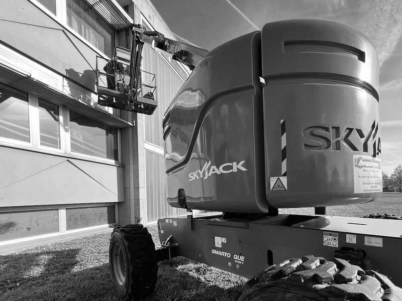Das Schwarz-Weiß-Bild zeigt eine große, geländegängige Arbeitsbühne der Marke "Skyjack" im Einsatz vor einem modernen Gebäude. Eine Person ist im Korb zu erkennen. Der klare Himmel und das Gebäude sind in kontrastreichen Grautönen dargestellt.