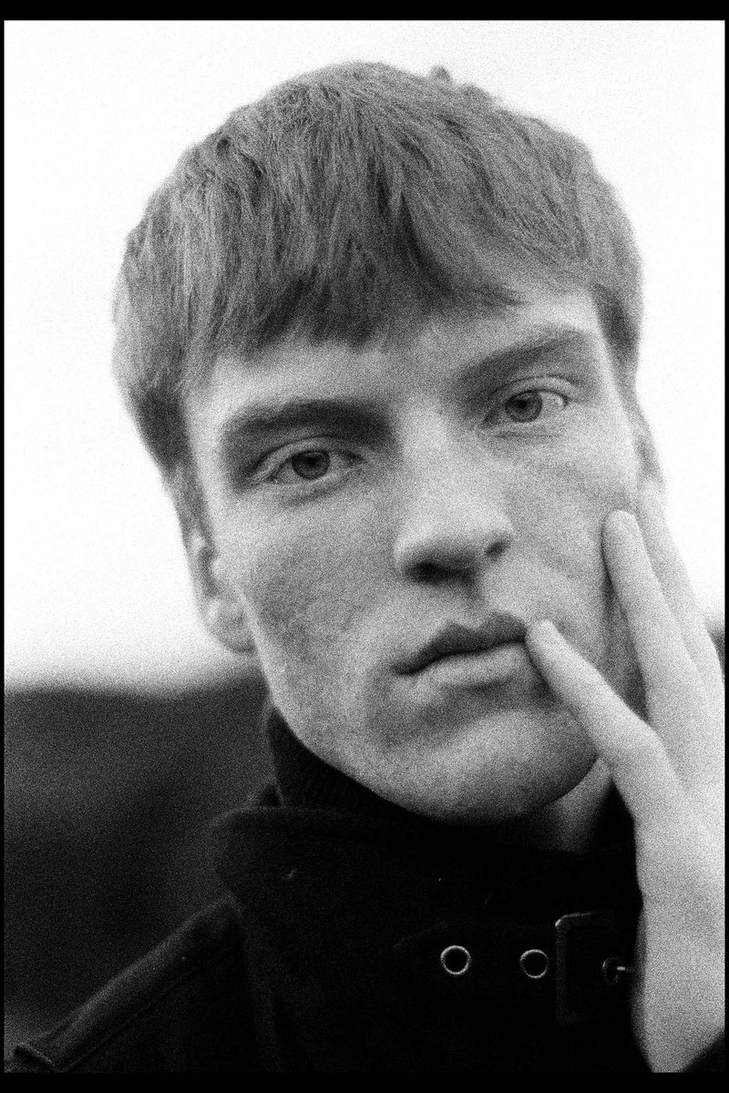 Black-and-white analog fine art portrait of a professional male model with short textured hair, hand resting against his cheek, wearing a dark high-collar jacket