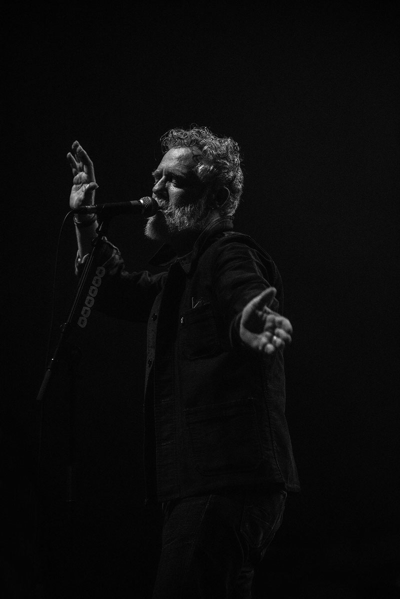 Black and white concert photograph of Glen Hansard singing into a microphone, both arms outstretched and hands gesturing expressively, performing at L'Olympia in Paris.
