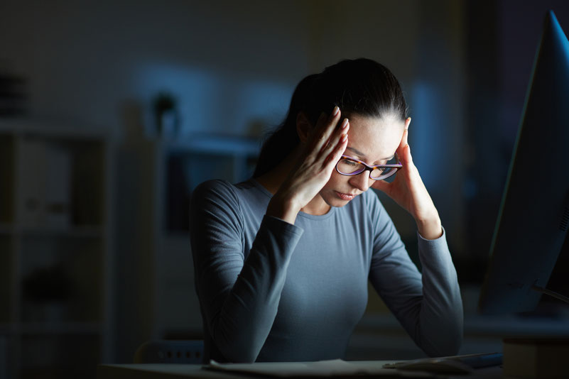 Eine Frau mit Brille sitzt nachts vor einem Computer und sieht gestresst aus, während sie sich mit beiden Händen an den Kopf fasst.