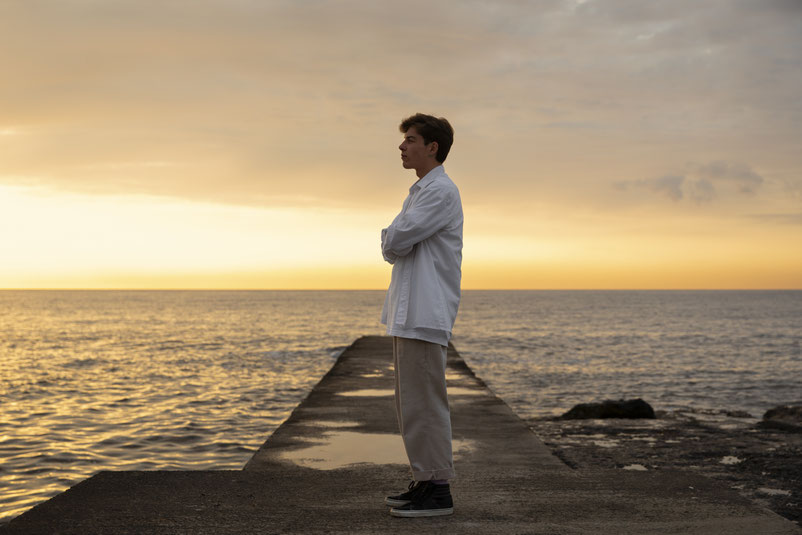 Ein junger Mann mit verschränkten Armen steht während eines goldenen Sonnenuntergangs auf einem Steg am Meer.