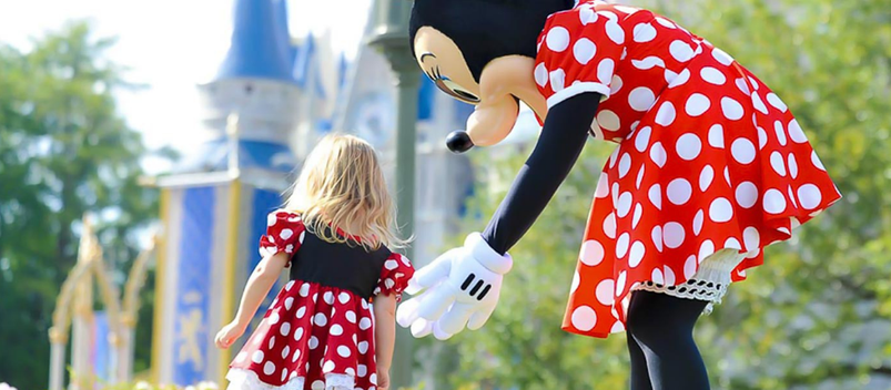 ディズニーワールドでミニーの衣装を着た子どもとキャラクターのミニーが向かい合う様子。後方にはシンデレラ城が見える。 (A child dressed in a Minnie costume interacts with Minnie Mouse character in front of Cinderella Castle at Disney World).
