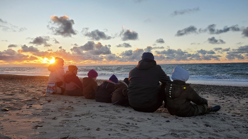 Eltern mit fünf Kindern sitzen bei Sonnenuntergang am Strand und blicken aufs Meer. Sind nur von hinten zu erkennen.
