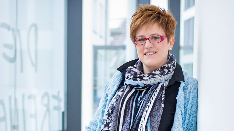 Julia Lakirdakis-Stefanou lehnt entspannt an einer Glaswand, freundlich lächelnd, mit roter Brille und gemustertem Schal – authentisch, kreativ und professionell bei juliaandfriends.de.
