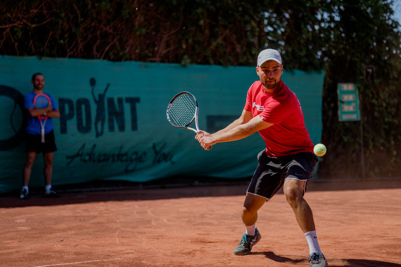 Zwei fortgeschrittene Tennisspieler von Ballplanet spielen auf einem Sandplatz in Magdeburg. Einer der Spieler trägt ein T-Shirt von Head und ist gerade dabei, eine kraftvolle Rückhand mit einem konzentrierten Blick auf den Tennisball auszuführen.