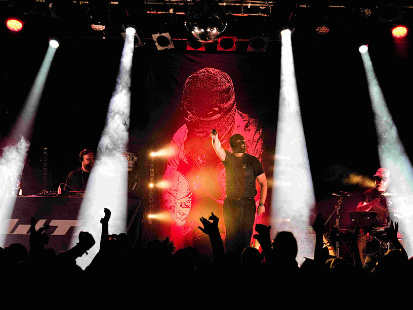 Live-Auftritt von Deutschrapper Waving the Guns auf der Bühne im Zakk Düsseldorf. Der maskierte Künstler in schwarzem Shirt rappt energiegeladen vor roter Leinwand mit seinem Porträt, begleitet von DJ am Pult. Bunte Bühnenlichter (rot, weiß, blau), Publik
