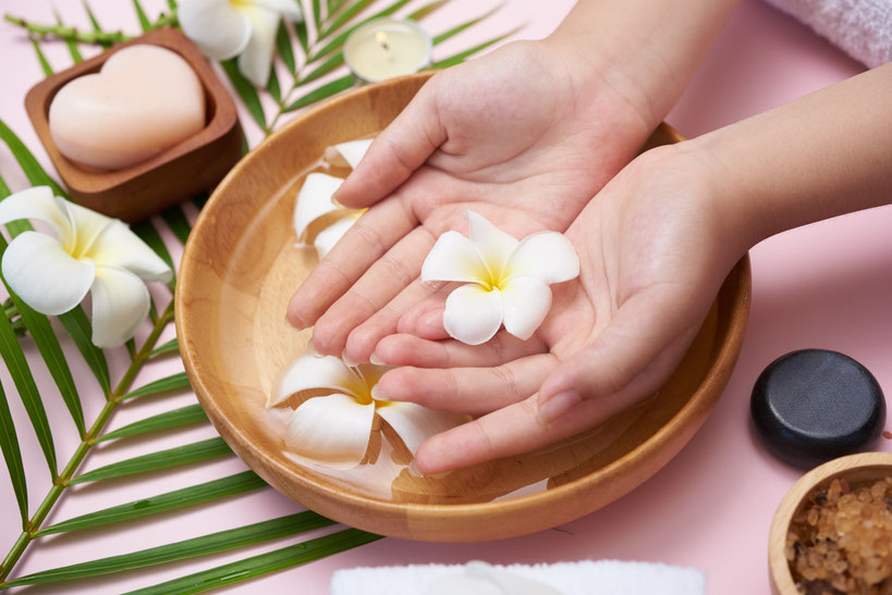 Hände halten eine weiße Plumeria-Blüte über eine Holzschale mit Wasser, umgeben von tropischen Blättern, einer herzförmigen Seife und Spa-Artikeln.