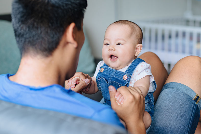 Ein fröhliches Baby sitzt auf dem Schoß des Vaters und schaut ihn erwartungsvoll an.