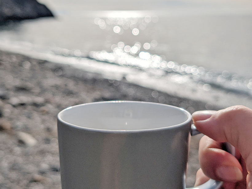 eine Hand hält eine graue Tasse Kaffee im Hintergrund sieht man verschwommen den Strand und das Meer