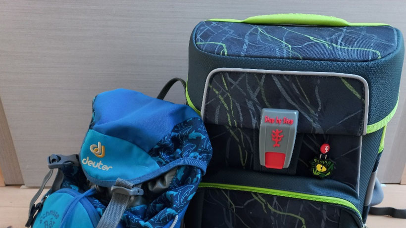 Blauer Kindergartenrucksack lehnt an der neuen Schultasche, vor einer grauen Tür.