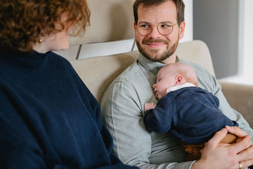 Vater mit schlafendem Baby auf der Brust sitzt neben seiner Frau auf dem Sofa.