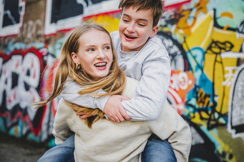 Schwester trägt lachend ihren Bruder Huckepack vor einer Graffitiwand.