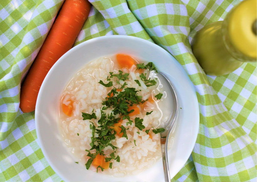 Mitte: weißer Teller mit Reisbrei, Möhren und Petersilie. Linke Ecke: Möhre. Rechte Ecke: grüner Pfefferstreuer. Untergrund: grün weiß karierte Tischdecke. Rezeptfoto.