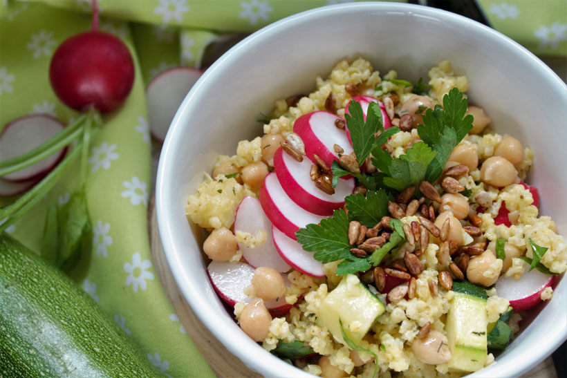 Rezeptfoto Low Carb Radieschen-Salat: Schüssel mit Hirse, Kichererbsen, Radieschen in Scheiben, Zucchini Würfel mit Petersilie. Neben der Schüssel liegt eine Zucchini und ein Radieschen mit grün.