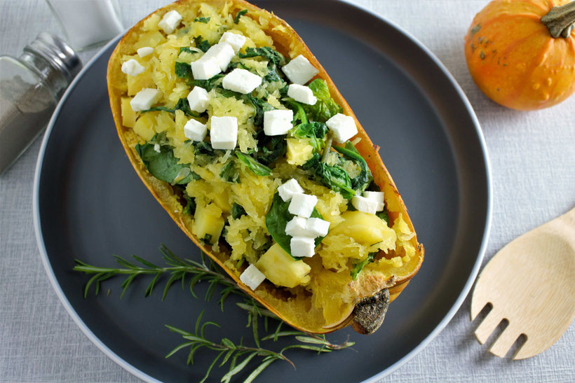 Mitte: grauer Teller mit einem Zweig Rosmarin. Darauf liegt ein halbierter Kürbis mit Spinat und Feta. Links oben: Salz und Pfefferstreuer. Rechts oben: Zierkürbis. Rechts unten: Kochlöffel. Rezeptfoto.