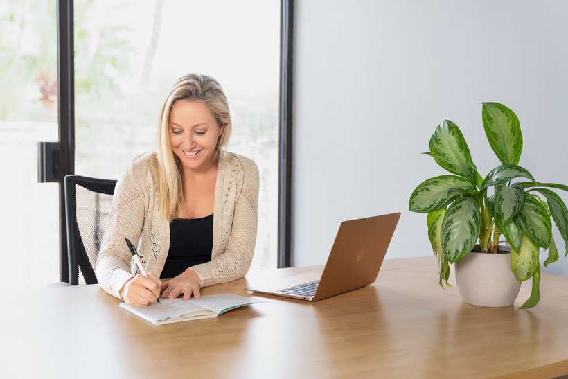 Woman at desk writing in calendar and smiling with laptop next to her and open