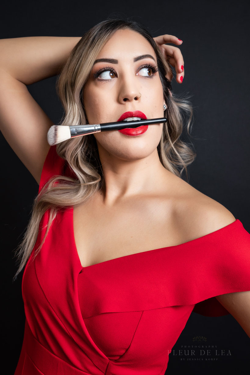 makeup artist woman with red lipstick and red jumpsuit holding makeup brush horizontally in teeth