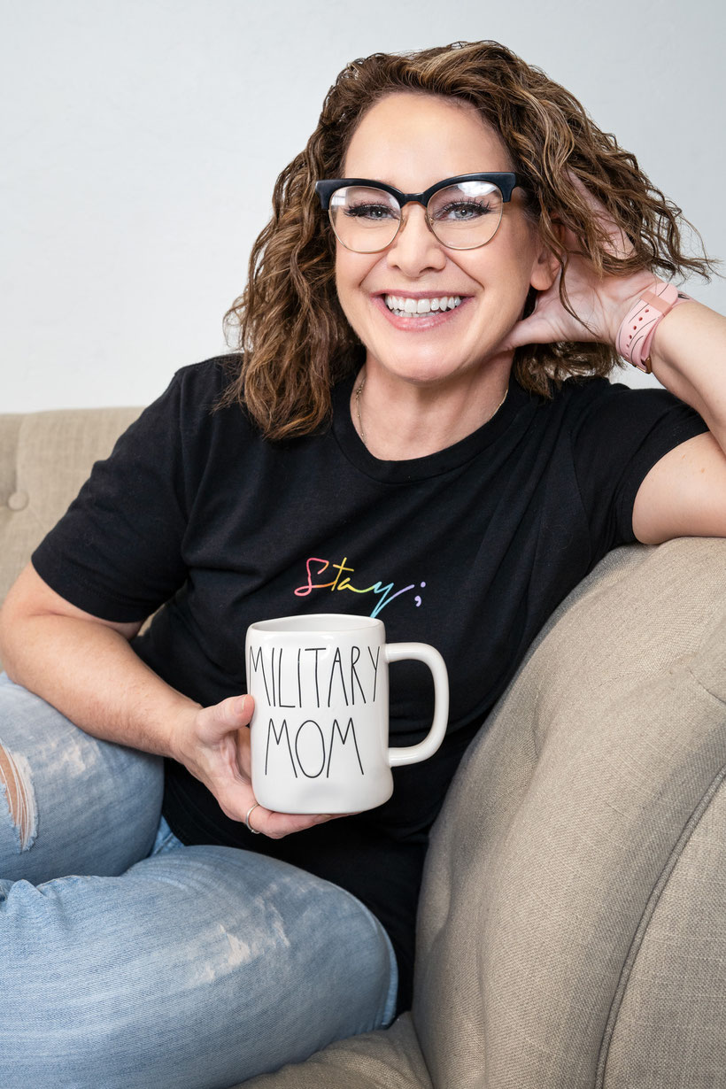 Woman leaning one arm on arm of couch, hand up behind ear. Holding a coffee cup that says Military Mom and wearing a shirt that says Stay in rainbow colors with a semi-colon