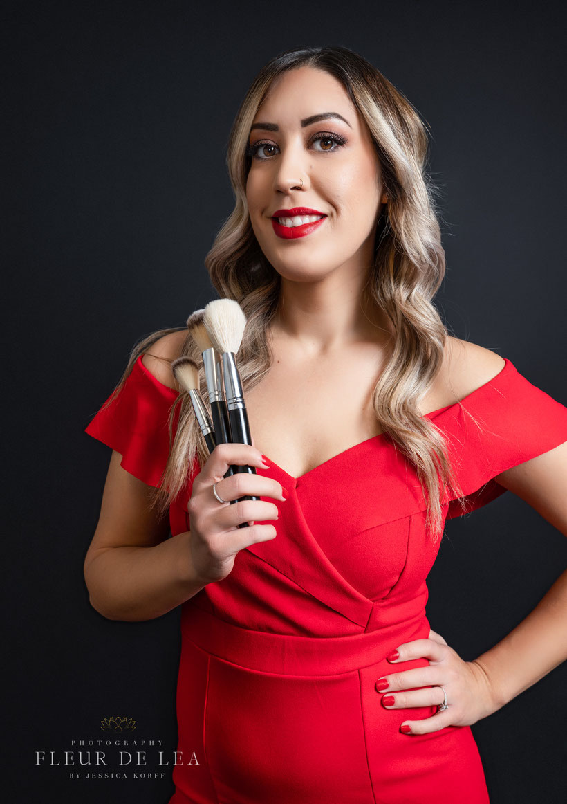 makeup artist woman holding brushes up and smiling with other hand on hip, wearing red jumpsuit and wearing red lipstick