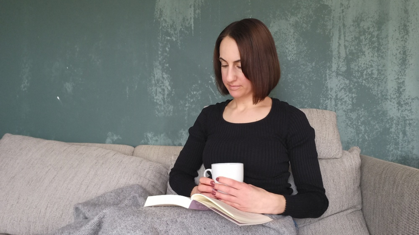 Michaela Dauscher sitzt auf einem Sofa, vor einen grünen Wand. Sie ist zugedeckt, liest ein Buch und hält eine Tasse in der Hand. Sie trägt ein schwarzes Oberteil.