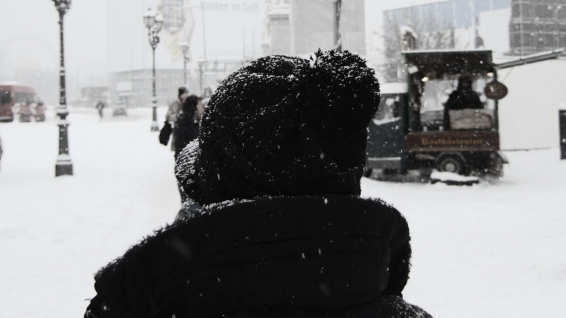 Nahaufnahme einer Person von hinten, es sind nur eine Strickmütze mit Bommel und der Kragen der Jacke zu erkennen. Es ist in schwarz weiß gehalten, es liegt Schnee und Straßenlaternen sind zu sehen. Auch ein kleiner Imbisswagen und ein paar Personen.