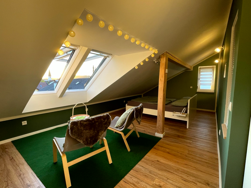 Schlafraum im FerienNest Kelheim mit Indoor-Loggia mit zwei Loungestühlen auf Rasenteppich unter großen Dachflächenfenstern, darüber Lichterkette, weißes Doppelbett hinter einem Holzpfosten, waldgrüne Wände