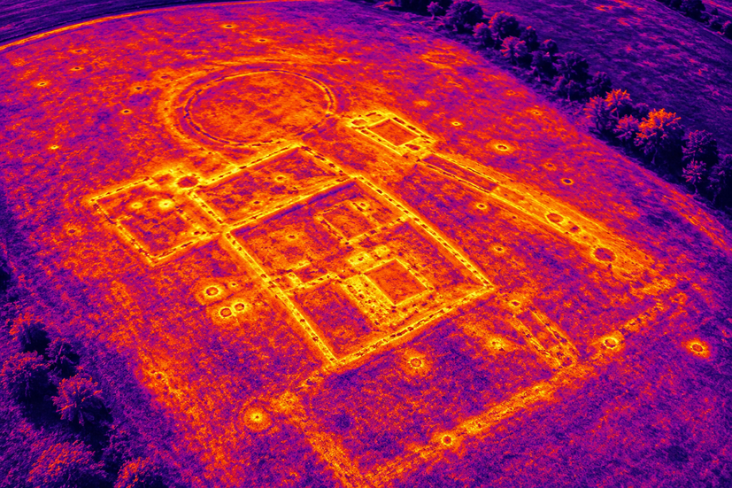 Thermografische Drohnenaufnahme einer archäologischen Fundstelle mit erkennbaren Bodenstrukturen und historischen Fundamentresten im Untergrund. 