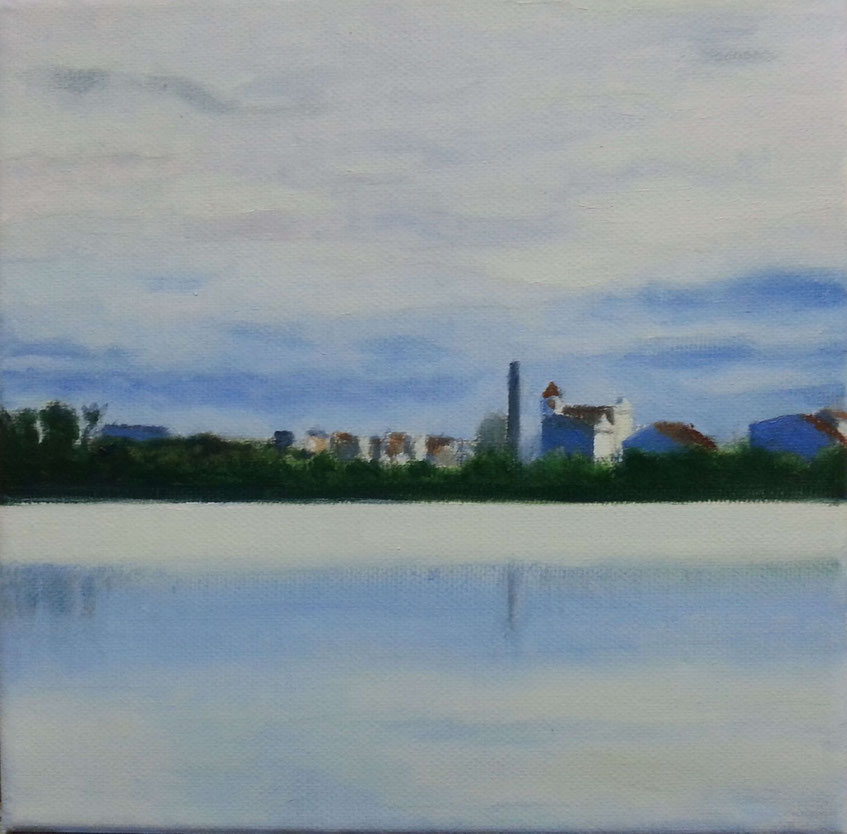 Construcciones de un molino junto a un cuerpo de agua en calma, con árboles y edificios al fondo bajo un cielo nublado.
