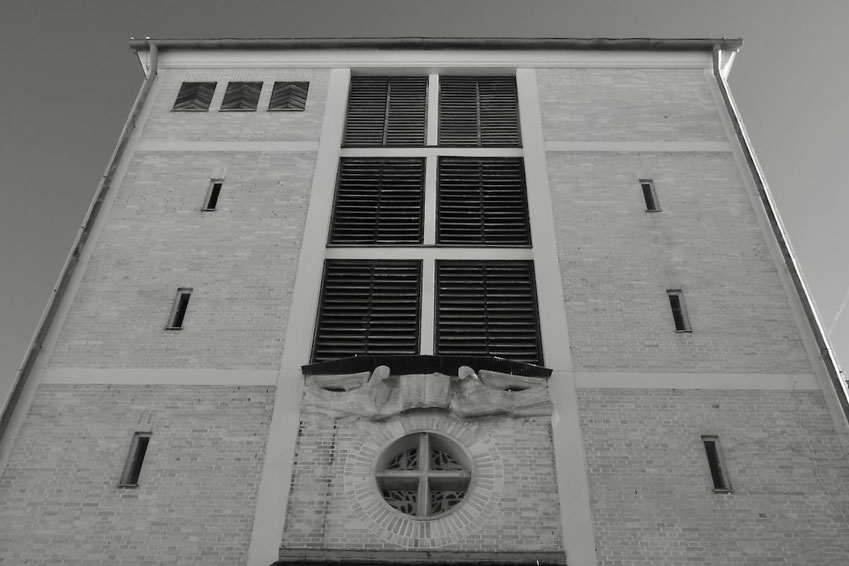 Architektur einer funktionalen Struktur in Schwarz-Weiß. Die Fassade zeigt eine Kombination aus massiven Backsteinwänden und großen, lamellenverkleideten Öffnungen.