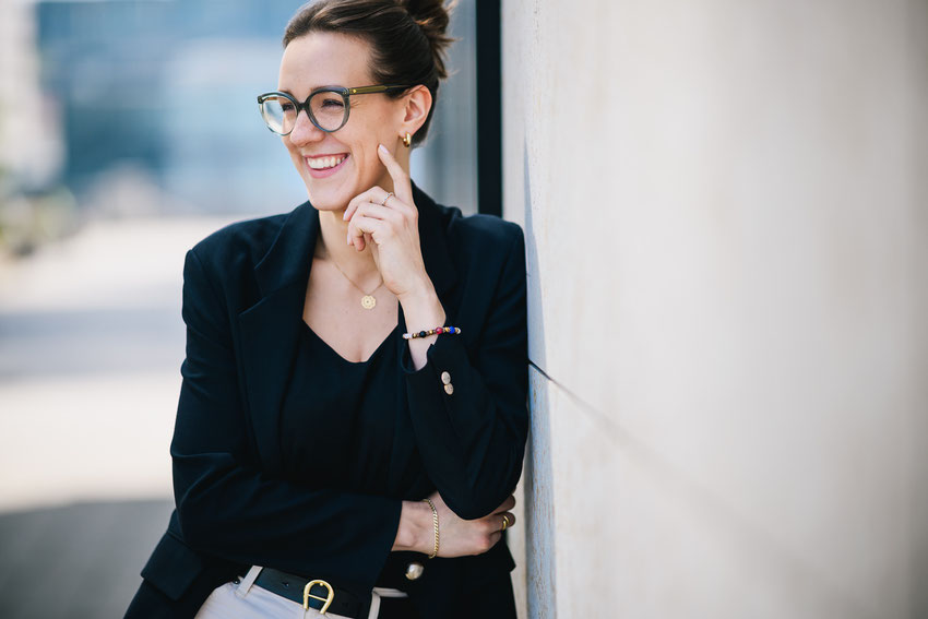 Portrait einer jungen Frau mit Brille und dunklem Blazer, die lächelnd zur Seite blickt und an einer Wand lehnt.