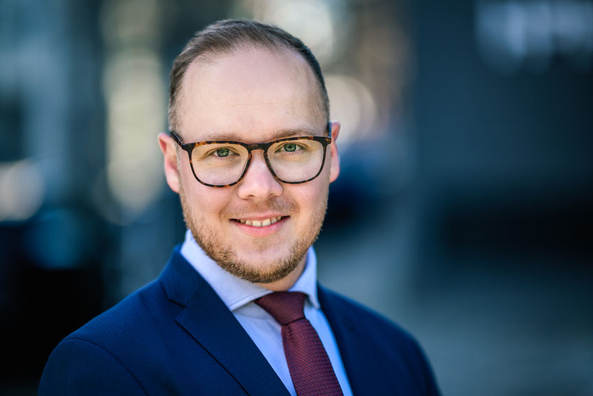 Lächelnder junger Mann mit Brille, Krawatte und blauem Anzug bei Tageslicht aufgenommen.