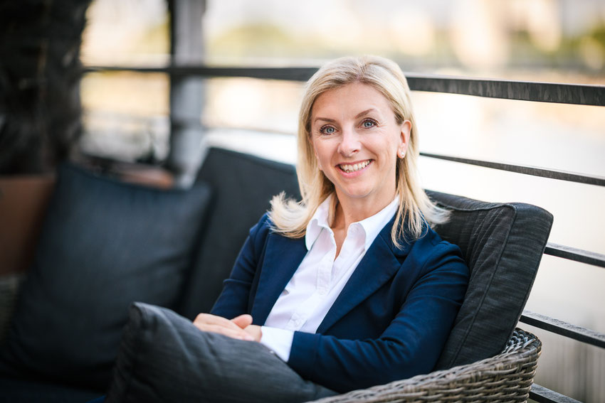 Freundliches Porträt einer sitzenden, blonden Frau in weißer Bluse und blauem Blazer.