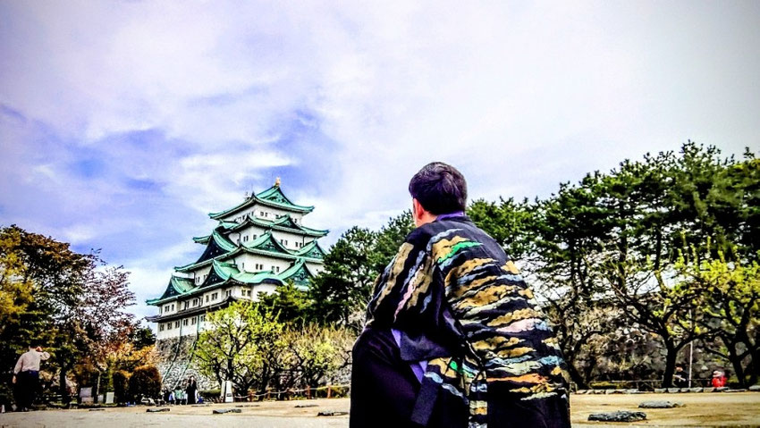 リラクゼーションサロン「forest hands（フォレストハンズ）」のセラピスト・徹平の背中越しから撮影した風景写真。左側には青空にそびえ立つ名古屋城の天守閣、右側手前には黒地に金、緑、水色、オレンジのグラデーションが入った波模様の豪華な羽織を羽織り、城を静かに見つめて佇む徹平の後ろ姿が写っている横長構図の画像。