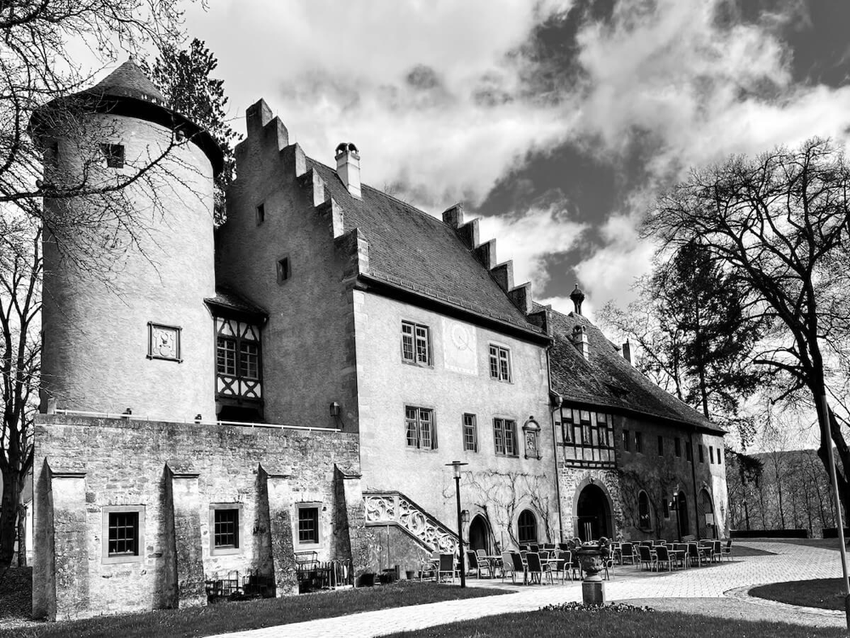 drei eng aneinandergebaute historische Gebäude eines Schlosses mit Turm, eingerahmt von alten Bäumen.