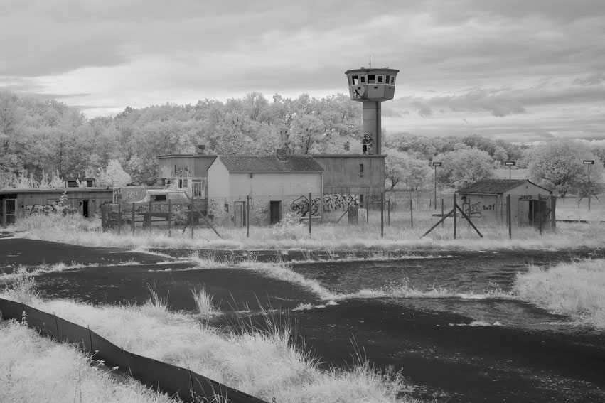 Infrarot-Foto eines verlassenen NATO-Geländes mit Wachturm, umzäunten Gebäuden und überwuchertem Asphalt vor dichtem Laubwald.