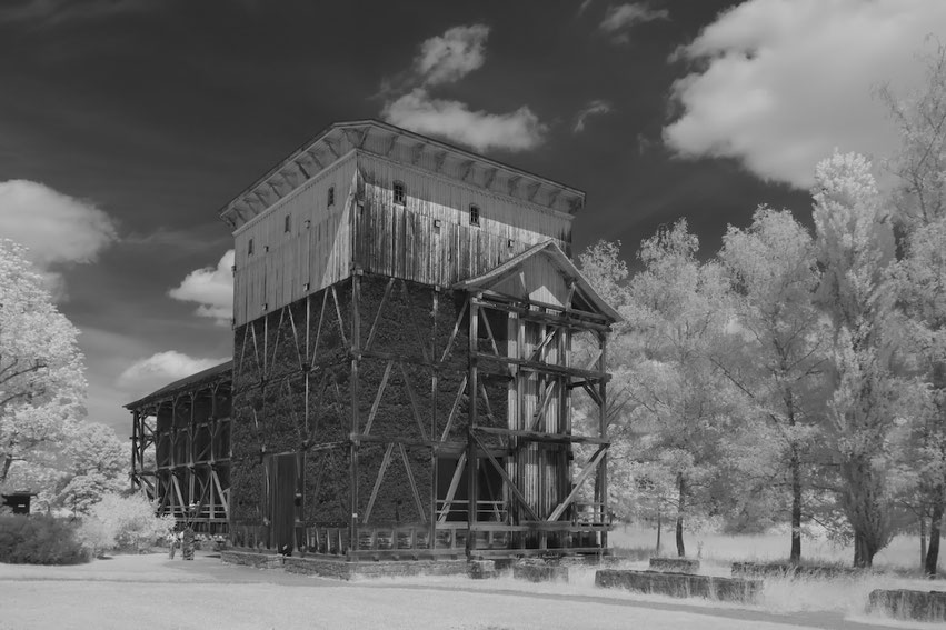 Das Gradierwerk in Bad Kissingen, ein markantes Bauwerk aus Holz und Reisig, erscheint in dieser Schwarz-Weiß-Infrarotaufnahme. Die umliegenden Bäume leuchten hell, was dem Bild eine besondere, fast ätherische Wirkung verleiht.