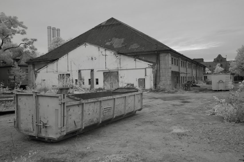 Infrarot-Foto einer zweigeschossigen, historischen Halle mit beschädigten Fenstern, losem Schutt im Vordergrund und Bauvorbereitung auf dem Gelände.
