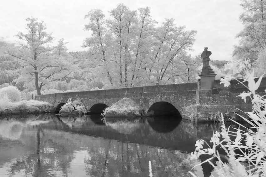 Eine historische Steinbrücke mit Bögen überquert die Sinn bei Schaippach. Eine Skulptur steht am Geländer, während die Bäume durch die Infrarotaufnahme in hellem Weiß erscheinen. Die Szene ist in Schwarz-Weiß gehalten, mit Spiegelungen im Wasser.