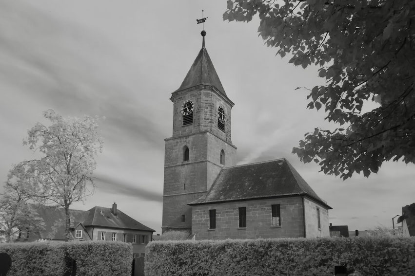 Die Evang.-Luth. Kirche St. Marien in Wallesau, Roth, mit ihrem markanten Turm ist in dieser Schwarz-Weiß-Infrarotaufnahme zu sehen. Die hell leuchtenden Bäume und die Hecke im Vordergrund verleihen der Szene eine besondere, leicht surreale Ästhetik.