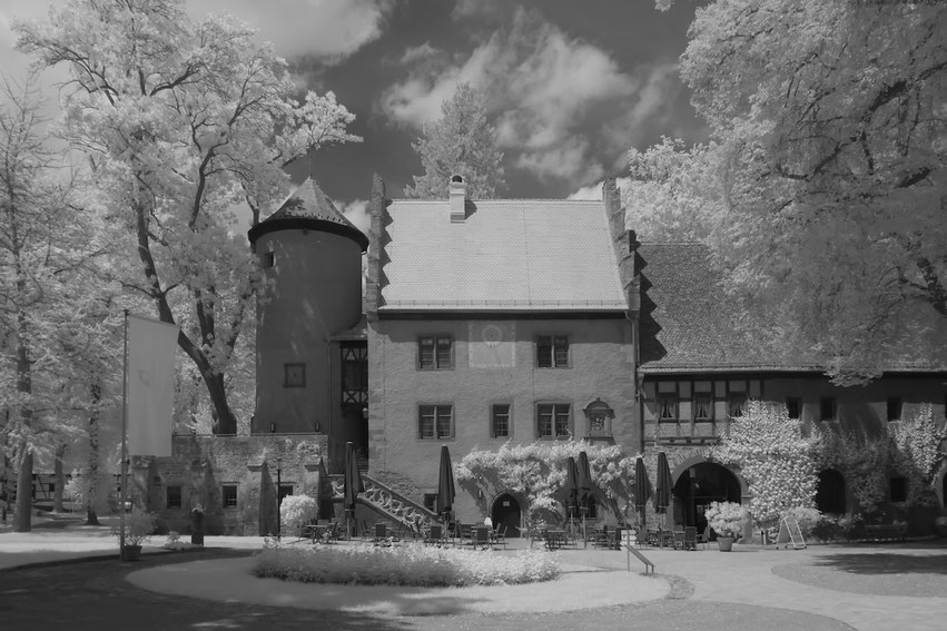 Das Gesindehaus von Schloss Aschach zeigt sich in Schwarz-Weiß mit Infraroteffekt. Das historische Gebäude ist umgeben von hell leuchtendem Baumbestand. Im Vordergrund sind Außenmöbel eines Cafés oder Museums zu sehen.