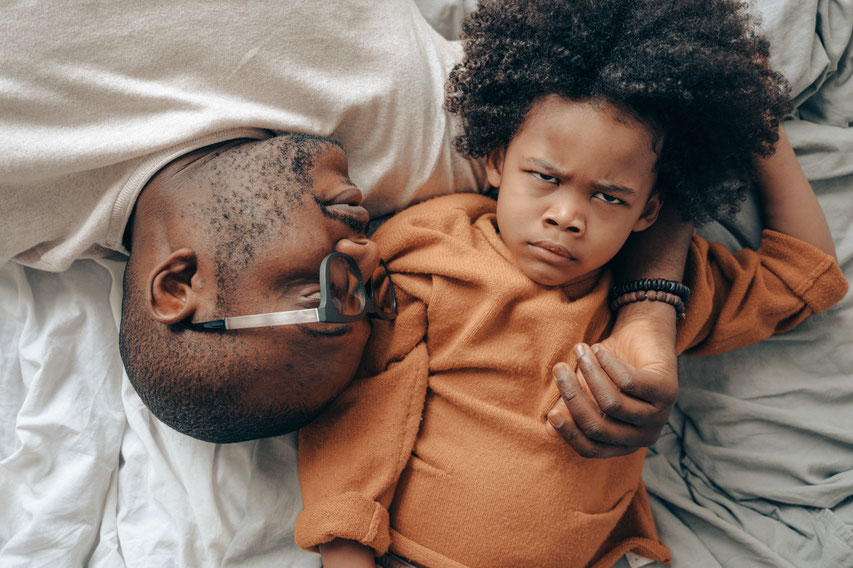 Vater und Sohn liegen auf dem Bett und ruhen sich aus nach dem Kindergarten, Das Kind istb wütend. Der Vater begleitet ihn liebevoll und bedürfnisorientiert
