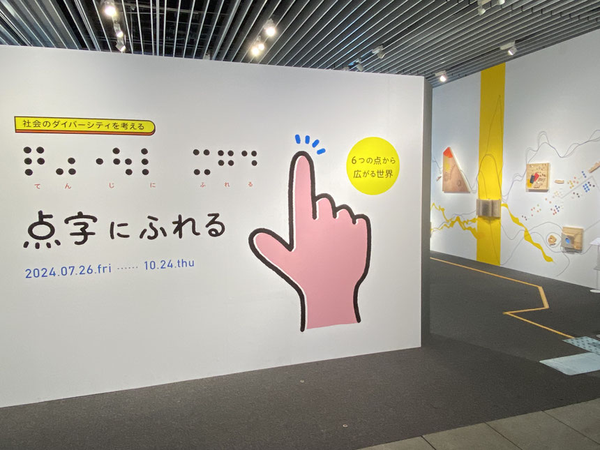 「点字にふれる」と点字でも墨字でも書かれている入口看板。点字に触ろうとするかのように人差し指を伸ばした手も描かれている。右手には壁があり展示作品が見える。床にはココテープが貼られている。